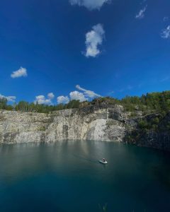 Image of “The Rock” in Quebec, Canada. A canoe and rider are featured in a bright lake with the cliff in the foreground, blue skies with a few white clouds.